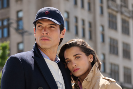 A man wearing a navy cap and coat stands next to a woman in a tan trench coat. Both look into the distance with a building in the background.